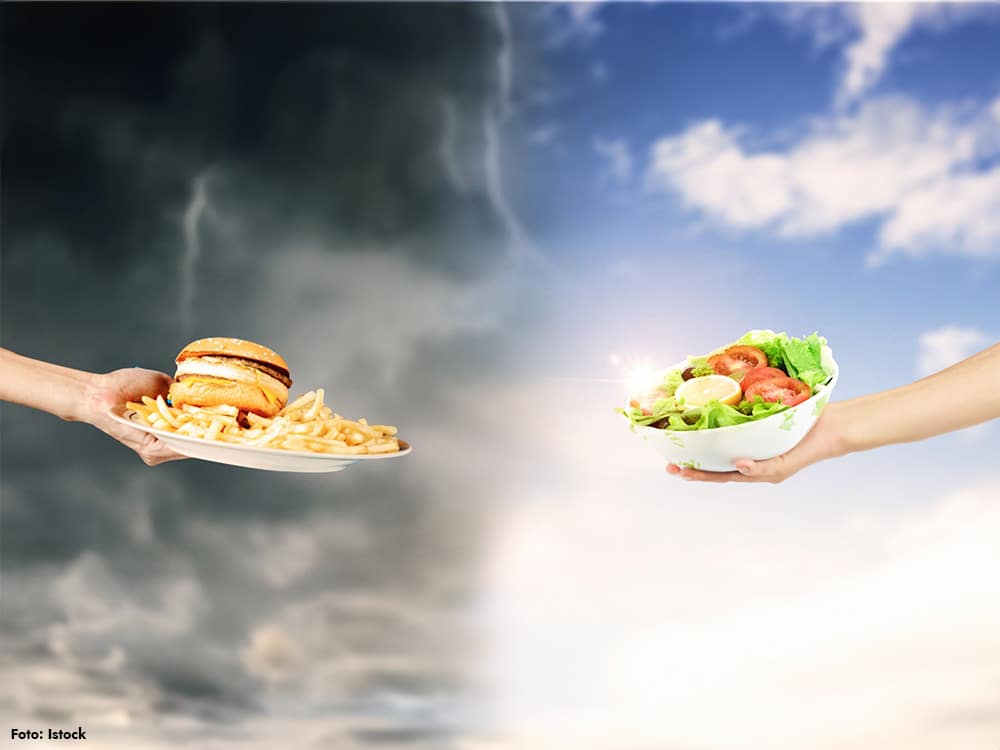 Illustration om ultraprocessad mat där en hand räcker fram hamburgare och pommes frites mot mörk himmel som exempel på ultraprocessad mat, och en annan hand räcker fram sallad som symbol för medelhavskost/medelhavsdiet – ett visuellt svar på frågan: vad är ultraprocessad mat?
