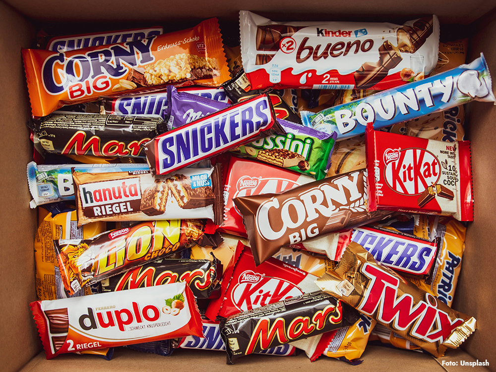 Kartong fylld med chokladkakor och godis (Snickers, Mars, KitKat m.fl.) – exempel på ultraprocessad mat och andra ultraprocessade livsmedel; illustrerar hur känner man igen ultraprocessad mat och kopplar till frågan vad är ultraprocessad mat.