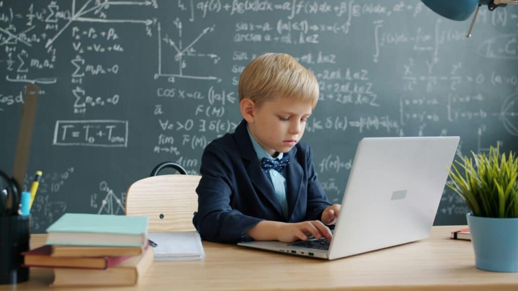 Varför särskilt begåvade barn inte alltid blir världsstjärnor som vuxna 2 Barn vid laptop i klassrum framför mattetavla med formler, illustration av särskilt begåvade barn och talangutveckling, medfödd talang och tidig specialisering.