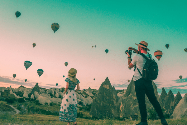 Resor och hälsa illustreras med två personer som fotograferar luftballonger över ett klipplandskap i solnedgången.