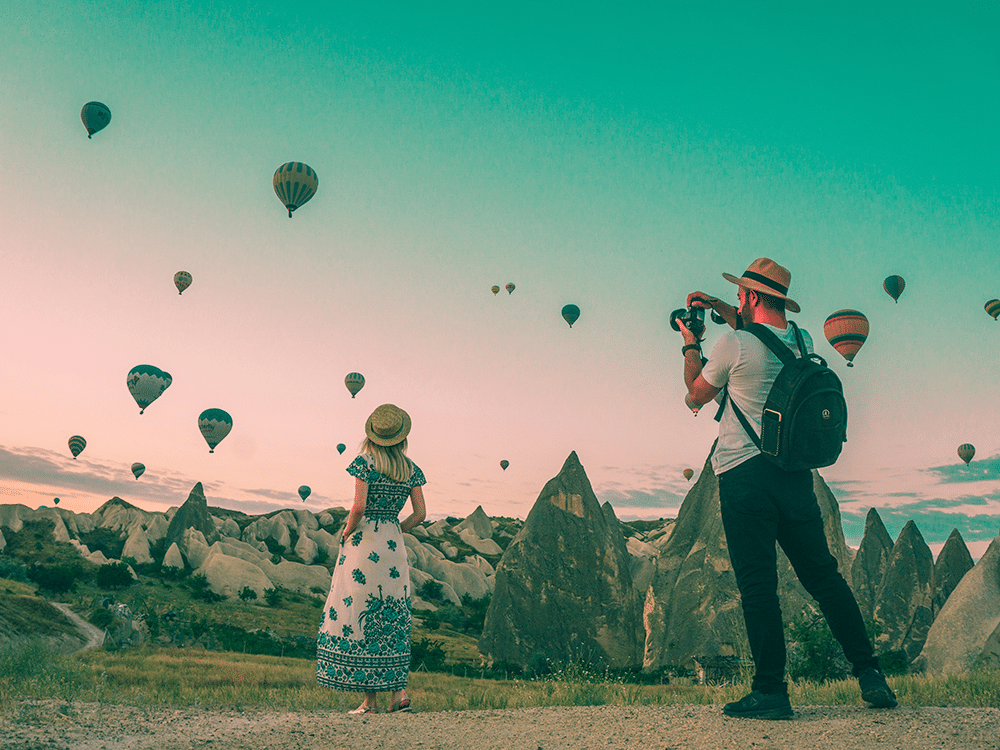 Resor och hälsa illustreras med två personer som fotograferar luftballonger över ett klipplandskap i solnedgången.