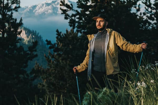 Man med gångstavar på en bergsstig omgiven av skog och höga berg.