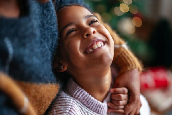 Leende barn som kramas av en vuxen framför julgransljus – varm bild av empati i vardagen.