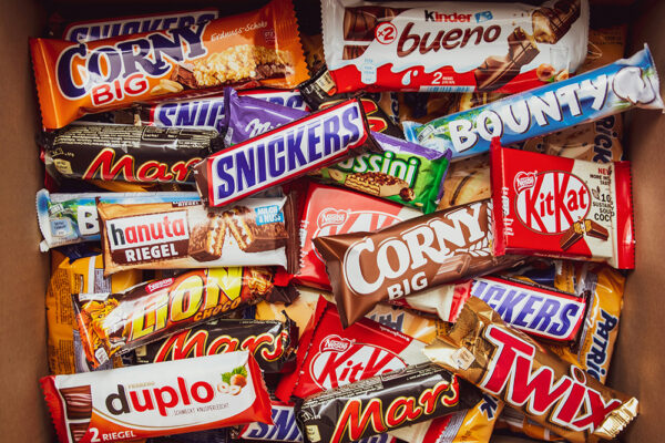Kartong fylld med chokladkakor och godis (Snickers, Mars, KitKat m.fl.) – exempel på ultraprocessad mat och andra ultraprocessade livsmedel; illustrerar hur känner man igen ultraprocessad mat och kopplar till frågan vad är ultraprocessad mat.