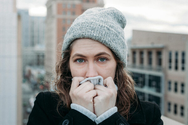 Svår förkylning: kvinna i mössa och kappa håller halsduken över munnen i kall stadsmiljö.